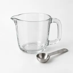 Centered clear glass measuring jug with a stainless-steel measuring spoon on a light neutral background.